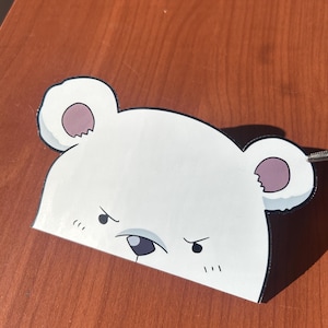 May include: A white, bear-shaped decorative item with black outlines and a stern expression. The bear has small, round ears with pink inner details and a black nose. The item is placed on a wooden surface.