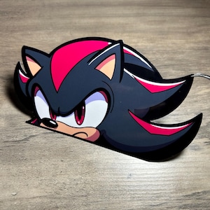 Puede incluir: Una pegatina troquelada con el personaje Shadow the Hedgehog de la serie Sonic the Hedgehog. La pegatina muestra la cara del personaje, con un cuerpo negro, detalles rojos y una expresión severa. Sobre una superficie de madera.