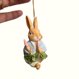 May include: A ceramic figurine of Peter Rabbit, wearing a blue jacket and holding two carrots. The rabbit has brown fur and is posed with a green lettuce skirt. The figurine is suspended by a gold string, suggesting it's an ornament.