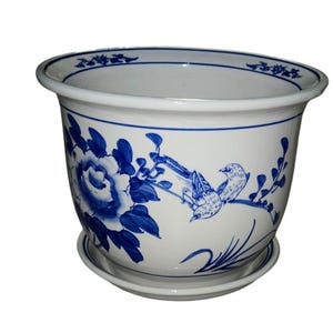 May include: A white ceramic planter with a matching saucer, featuring a blue hand-painted design of a large flower and two birds perched on a branch. The rim of the planter is decorated with a thin blue line and small floral motifs.