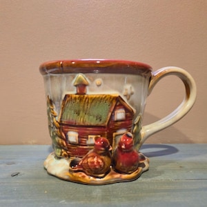 May include: Ceramic mug with a winter scene featuring a red cabin with a green roof, trees, and two red birds. The mug has a brown rim and a cream-colored handle. The base of the mug is sculpted with a decorative design.