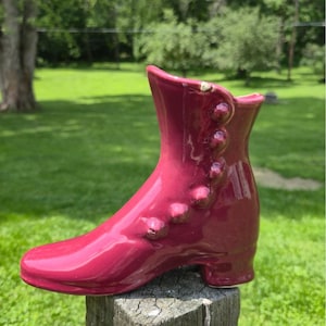 May include: A burgundy ceramic boot-shaped vase. The vase has a glossy finish and features a row of decorative buttons along the side. The boot is set on a wooden post against a backdrop of green grass and trees.
