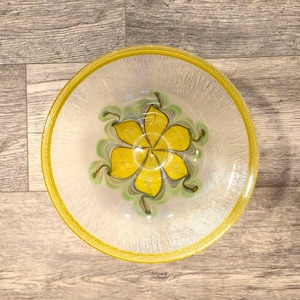 May include: A round glass bowl featuring a vibrant yellow flower design at the center with swirling green accents. The bowl has a translucent, textured body and a solid yellow rim, creating a clear visual contrast between the center and edges.
