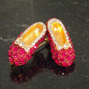 May include: A pair of miniature shoe-shaped brooches covered in vibrant fuchsia crystals. Each piece features a gold-tone interior, a clear crystal-encrusted trim, and a small bow detail at the toe, creating a sparkling, decorative accessory set.