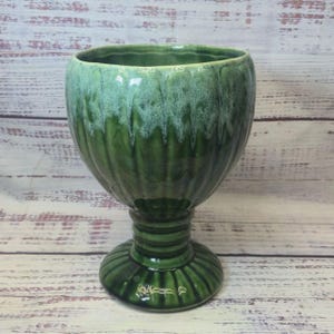 May include: A green ceramic goblet with a ribbed design. The goblet has a wide bowl and a pedestal base. The glaze has a dripped effect, with lighter green and white accents at the top. The goblet is on a light wood surface.