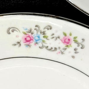May include: A white ceramic plate featuring a delicate floral pattern along the rim. The design includes small pink and blue roses accented by grey swirling leaves and stems. The edge of the plate is finished with a thin silver metallic trim.