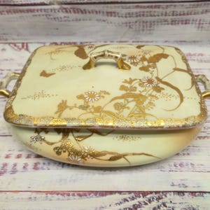 May include: A rectangular porcelain serving dish with a lid, featuring a cream-colored base decorated with gold floral patterns, delicate white daisy-like flowers, and a butterfly. The dish includes two gold-toned handles and a central lid handle.