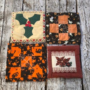 May include: Four square quilted pot holders. One features a holly design, another has a Halloween theme with ghosts and moons, a third has black cats and bats, and the last has a poinsettia design.