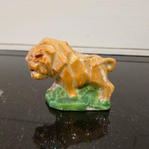May include: A small, ceramic lion figurine in shades of orange and brown, with a green base suggesting grass. The lion has a geometric, angular design and is positioned in a side profile. The figurine is likely a decorative item.