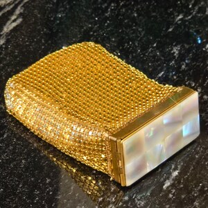 May include: A rectangular gold-toned metal mesh case featuring a hinged lid adorned with a grid of iridescent white mother-of-pearl squares. The body of the case is constructed from a fine, flexible metallic mesh with a textured surface.
