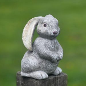 May include: A gray bunny statue with long ears and a textured surface. The bunny is sitting upright with its paws together. The statue is placed on a wooden post against a blurred green background. A decorative garden figurine.