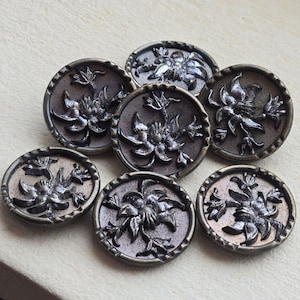 May include: Seven antique metal buttons with a floral design. Each button features a raised, detailed flower in the center, surrounded by a textured border. The buttons have a tarnished, aged appearance, with shades of gray and bronze.