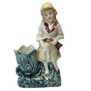 May include: A porcelain figurine of a girl wearing a straw hat and holding a doll. She is standing on a green and gold base with a flower pot.