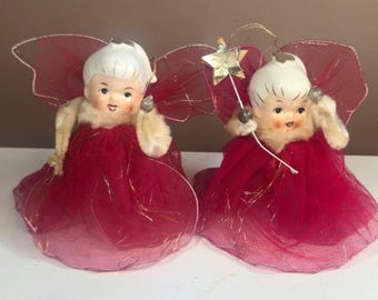 May include: Two vintage-style angel ornaments featuring white ceramic faces with painted features, white hair, and red tulle dresses with gold trim. Each angel has sheer red wings and one holds a gold star wand. The figures have fuzzy white arms.