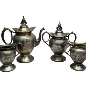 May include: A four-piece silver-toned tea set consisting of a large teapot with a lid, a lidded sugar bowl with two handles, a creamer, and a waste bowl. Each piece features an ornate engraved pattern, a pedestal base, and a tarnished metallic finish.