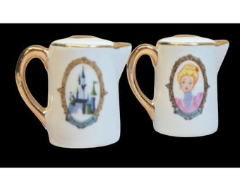 May include: Two white ceramic creamer pitchers with gold trim. One features a colorful illustration of Cinderella's castle, the other features a portrait of Cinderella. Both have the text "Walt Disney World" on the bottom.