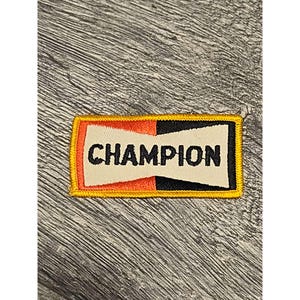 May include: A rectangular patch with the word "CHAMPION" in black letters. The patch has a white center, with orange and black sections on either side. A yellow border surrounds the patch.