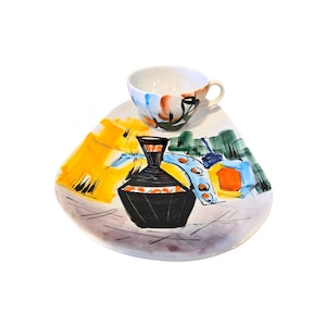May include: A white ceramic plate with a colorful abstract design featuring a black vase with orange accents. A white ceramic cup with a floral design sits on top of the plate.