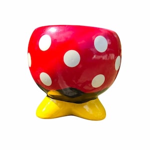 May include: A red ceramic planter with white polka dots. The planter has a yellow base and a black band. The planter is designed to resemble a cartoon character.