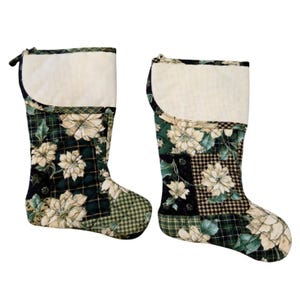 May include: Two quilted Christmas stockings featuring a patchwork design of cream-colored poinsettia flowers on dark green and black plaid backgrounds. Each stocking has a solid cream-colored cuff at the top and a hanging loop on the side.