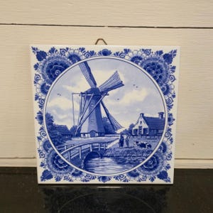 May include: Square ceramic tile with a blue and white windmill scene. The windmill is centered in a circular design, surrounded by a floral border. A bridge, house, and cows are also depicted. The tile is likely for decorative purposes.