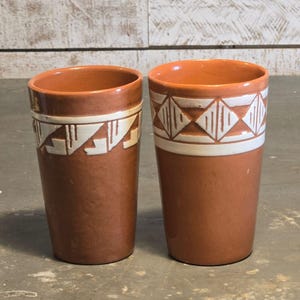 May include: Two terracotta-colored ceramic tumblers featuring white painted geometric patterns around the upper portion. The left tumbler displays a stepped design, while the right tumbler features a repeating diamond pattern. Both vessels are cylindrical.