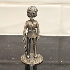 May include: A small, metallic figurine of a British Royal Guard in a traditional uniform. The figure is detailed with a tall, fuzzy hat, a buttoned jacket, and a rifle. The figurine stands on a textured, circular base, and is a bronze color.