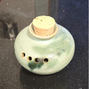 May include: A round, light green ceramic container with a cork stopper. The container has three small holes on the side and a darker green mottled pattern. The cork is light brown.