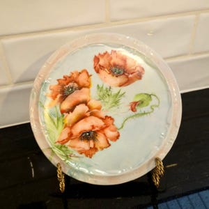 May include: A round decorative plate featuring a watercolor painting of orange poppies with green leaves and buds. The plate has a light blue center and a pink rim. It is displayed on a gold-colored metal stand.