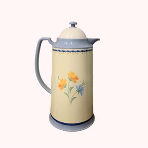 May include: A vintage cream-colored coffee carafe with a light blue handle, lid, and base. The carafe features a floral design with orange and blue flowers. The carafe is approximately 12 inches tall.