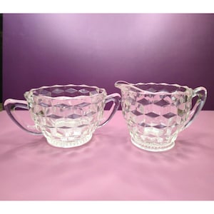 May include: A clear glass sugar bowl and creamer set. Both pieces have a diamond pattern and scalloped edges. The sugar bowl has two handles. The creamer has a spout and a single handle. The set is on a pink surface with a purple background.
