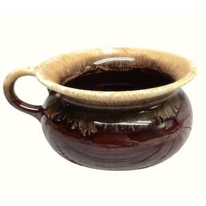 May include: A round ceramic bowl with a single handle, featuring a deep dark brown base color that transitions into a lighter tan and cream drip glaze pattern around the wide, flared rim and the upper exterior section of the vessel.