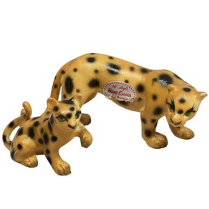 May include: A pair of bone china leopard figurines featuring a larger standing leopard and a smaller crouching leopard. Both figures have a tan base coat with black spots, green eyes, and a sticker on the larger leopard reading BONE CHINA.