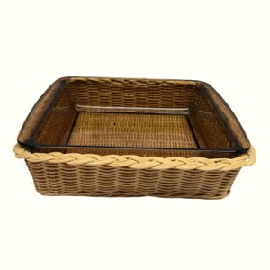 May include: A square, transparent amber-colored glass baking dish nested inside a light beige woven wicker basket holder. The basket features a braided rim detail around the top edge, providing a secure fit for the glass container.