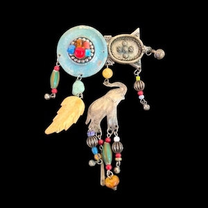 May include: A vintage-style brooch with a light blue circular base, adorned with colorful stones and a silver elephant charm. The brooch features dangling beads in various colors and shapes, and a carved leaf. The background is black.