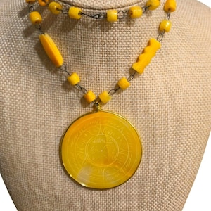 May include: A yellow necklace with a large, round pendant. The pendant has a gold-colored frame and a detailed, etched design. The necklace features yellow beads and a dark chain. The necklace is displayed on a beige mannequin.