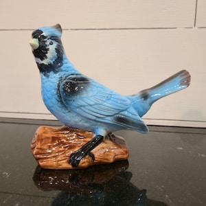 May include: A ceramic figurine of a blue jay perched on a brown log. The bird has light blue feathers with black accents on its wings and head. The beak is yellow, and the bird's feet are black. The figurine is a decorative item.