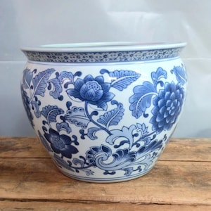 May include: A round ceramic planter featuring a white base with intricate blue floral patterns and scrolling leaf designs. The rim is decorated with a thin, repeating geometric border pattern, and the overall shape is wide and bulbous.