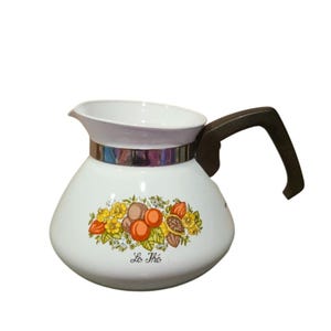 May include: White vintage teapot with a brown handle and a silver band. The teapot features a floral design with the text "Le Thé" in black. The teapot is designed for serving tea.