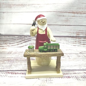 May include: A snowman figurine wearing a red Santa hat and apron, holding a hammer and working on a green toy truck on a wooden workbench.