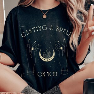 Puede incluir: Una camiseta negra con un gráfico de dos manos lanzando un hechizo con una luna creciente y cristales. El texto en la camiseta dice "Casting a spell on you".