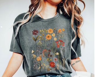 Puede incluir: Una camiseta gris con un estampado floral en varios colores, incluyendo rojo, naranja, amarillo y verde. Las flores están dispuestas en un grupo en la parte delantera de la camiseta.