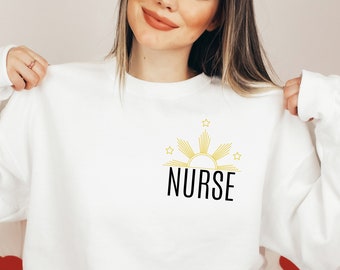 May include: White sweatshirt with a yellow sun and stars graphic and the word "NURSE" in black text.