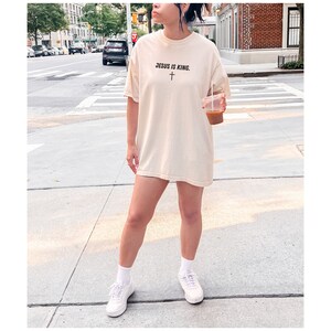 K&ouml;nnte beinhalten: Cremefarbenes T-Shirt mit dem Text "JESUS IS KING" und einer schwarzen Kreuzgrafik. Das Shirt wird mit wei&szlig;en Turnschuhen und Socken getragen. Das Bild wird im Freien auf einer Stadtstra&szlig;e aufgenommen.