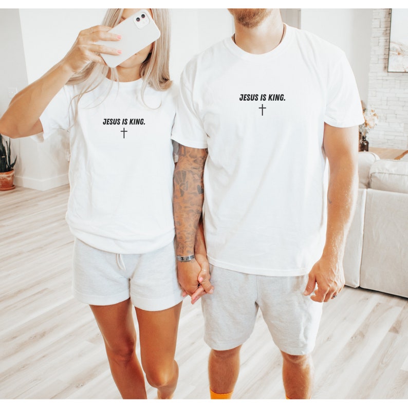 K&ouml;nnte beinhalten: Zwei Personen tragen passende wei&szlig;e T-Shirts und graue Shorts. Die Shirts tragen den Text "JESUS IS KING" &uuml;ber einem kleinen Kreuz. Sie halten H&auml;ndchen. Der Hintergrund ist ein Raum in neutralen Farbt&ouml;nen.