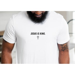 K&ouml;nnte beinhalten: Wei&szlig;es T-Shirt mit dem Text "JESUS IS KING." in Schwarz, zusammen mit einem schwarzen Kreuzsymbol. Das Shirt besteht aus einem weichen Material und ist f&uuml;r den t&auml;glichen Gebrauch geeignet.