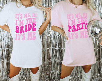 Puede incluir: Dos camisetas blancas con texto rosa. La primera camiseta dice "It's me, hi I'm the bride it's me". La segunda camiseta dice "It's me, hi I'm the party it's me".