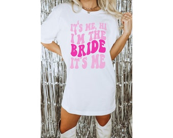 Puede incluir: Camiseta blanca con texto rosa y blanco que dice "It's me, hi I'm the bride it's me" en una fuente retro.