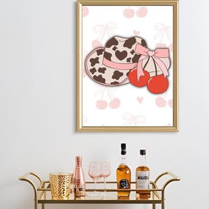 May include: A gold bar cart with two glass shelves holds bottles, glasses, and a decorative candle holder. A framed print above features a cowboy hat with a pink bow and cherries.