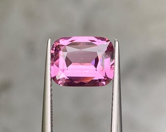 May include: A faceted, rectangular pink gemstone held by silver tweezers. The gem has a vibrant, light pink hue with multiple facets that reflect light. The gemstone is a rectangular cushion cut.